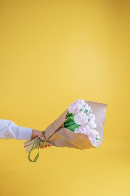 A person in a white shirt holds a bouquet of fresh, pale pink tulips wrapped in light brown paper and tied with a green ribbon against a solid yellow background. The tulips have smooth, slightly curve