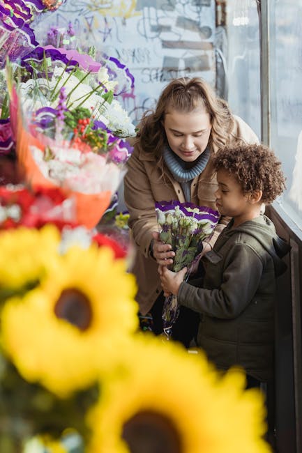 A woman with wavy, shoulder-length hair and a light blue turtleneck sweater, smiling softly, presents a bouquet of purple and white flowers wrapped in clear plastic to a young boy with curly brown hai