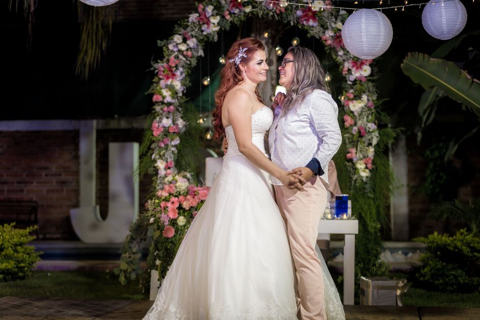 A wedding celebration features a bride dressed in a strapless white gown with lace detailing, holding hands with a woman in a light-colored blazer and pink trousers. They are standing outdoors under s