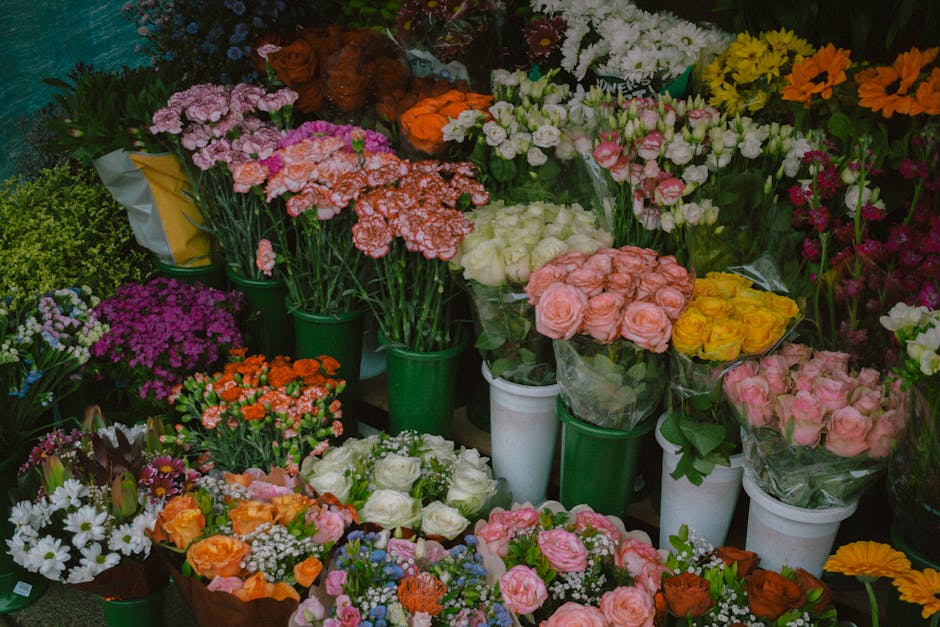 A vibrant display of floral arrangements with various fresh flowers including pink, yellow, orange, white, and purple blossoms. The flowers are arranged in bouquets wrapped in clear or plain paper, pl