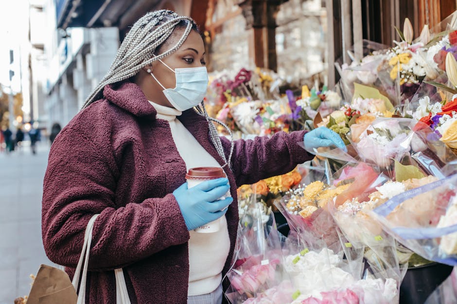 A woman wearing a face mask and blue gloves is selecting a bouquet of fresh flowers from an outdoor floral stall on Abbey Wood High Street. She is holding a takeaway coffee cup in her other hand and i