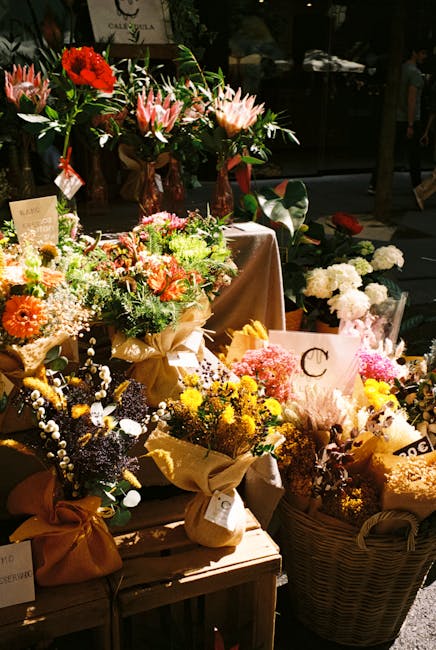 A display of various floral arrangements featuring a vibrant mix of fresh flowers, including red and pink proteas, white and yellow chrysanthemums, and sprigs of white baby's breath, all set on a wood